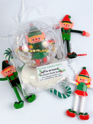 Bendable elf treasure toy bath bomb, clear ornament with white/gold crinkle paper, on top of a white bath bomb inside a clear box.